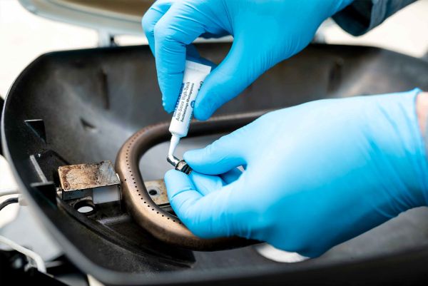 A person wearing blue gloves applies anti-seize paste from a tube onto a small metallic screw inside a grill housing.
