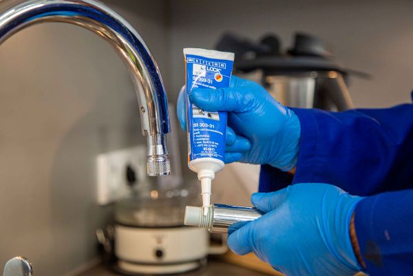 A person wearing blue gloves applies WEICON thread sealant from a tube onto the threaded fitting of a faucet component.
