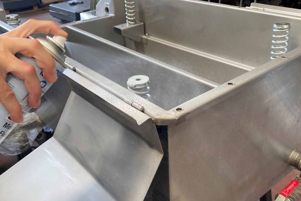 A hand sprays product from an aerosol can onto a stainless steel industrial container joint.
