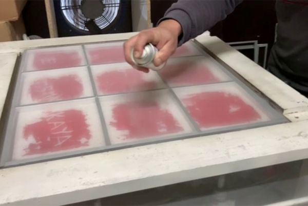 A person applies spray adhesive from a can onto a screen printing frame grid.