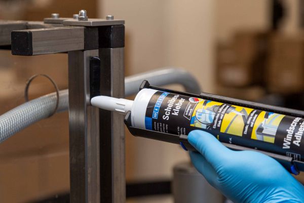 A person wearing a blue glove applies WEICON Windscreen Adhesive from a dispensing gun onto an industrial metal frame.