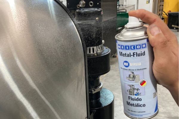 A hand sprays WEICON Metal-Fluid from a spray can onto a brushed stainless steel surface of an industrial machine.