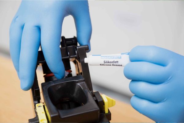 A person wearing blue gloves applies WEICON Silicone Grease from a small tube onto a component of a mechanical assembly.