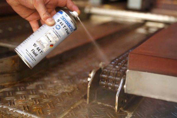 A hand sprays WEICON W 44 T-Fluid onto an industrial drive chain component.