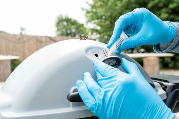 A person wearing blue gloves applies anti-seize paste from a tube onto the thread of a temperature gauge being installed on a grill lid.