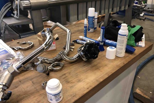 Stainless steel pipes, pipe clamps, and various WEICON spray cans, sealants, and containers are arranged on a wooden workbench.